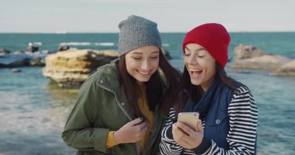 Dois belos melhores amigos do sexo feminino em chapéus de pé na praia e rir ao discutir foto no smartphone. — Vídeo de Stock