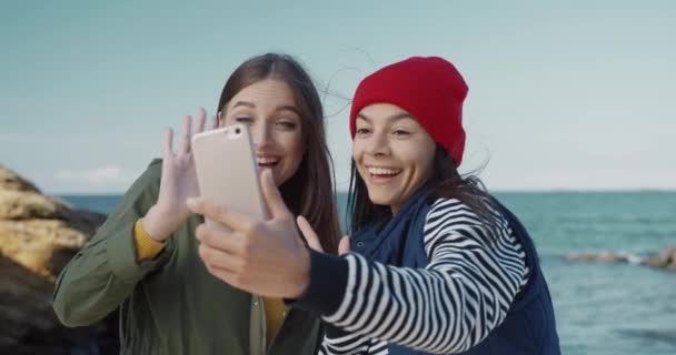 Dos hermosas chicas jóvenes están de pie junto al mar y hablando en videollamada. Felices amigas viajan juntas y filman blog. — Vídeos de Stock