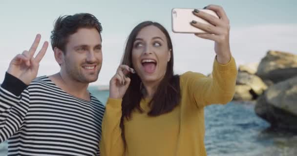 Boldog fiatal pár grimaszok tartása okostelefon a kezében, és hogy vidám fotó hátterében a tenger és a hullámok. — Stock videók