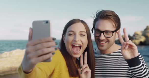 Šťastný mladý pár se fotí na pozadí moře a kamení. Fešák chlap a dívka, aby selfie zatímco stojí na břehu. — Stock video