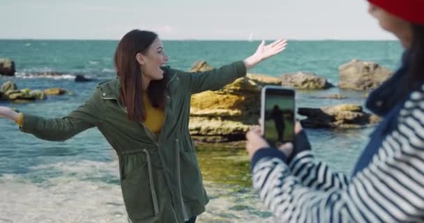 Hermosa chica caucásica en chaqueta elegante se regocija y gira sonriendo en la orilla del mar mientras su amigo está filmando en el teléfono. — Vídeos de Stock