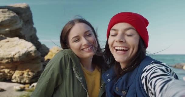 Close-up de duas jovens garotas alegres se divertindo posando na câmera do smartphone enquanto estão na costa do mar no outono. Vista da câmara. — Vídeo de Stock