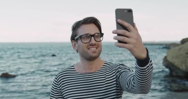 Fešák běloch v brýlích se sněhobílým úsměvem drží smartphone mluví na video volání na pozadí moře. — Stock video