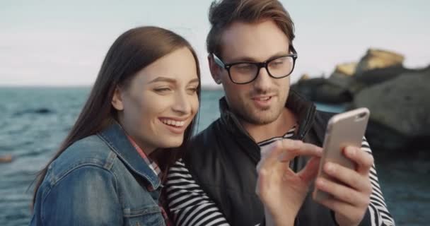 Joven pareja feliz de pie junto al mar revisando y discutiendo fotos en el teléfono inteligente. Guy magnifica imagen con gesto en pantalla. — Vídeos de Stock