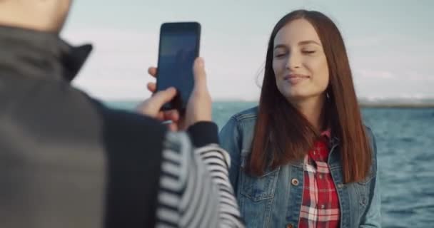 Krásná běloška dívka v džínové bundě s tekoucími vlasy víry a pózuje pro kameru, zatímco stojí na pláži, když přítel fotí ji. — Stock video