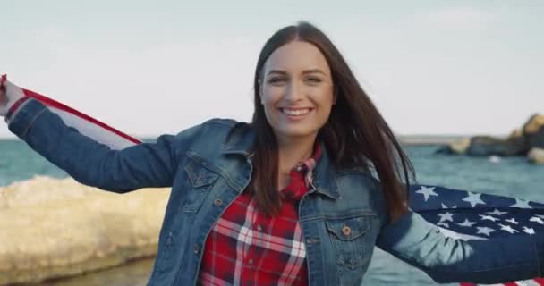 Närbild porträtt av glad kvinnlig student flicka står mot bakgrund av havsvågor insvept i flagga USA — Stockvideo