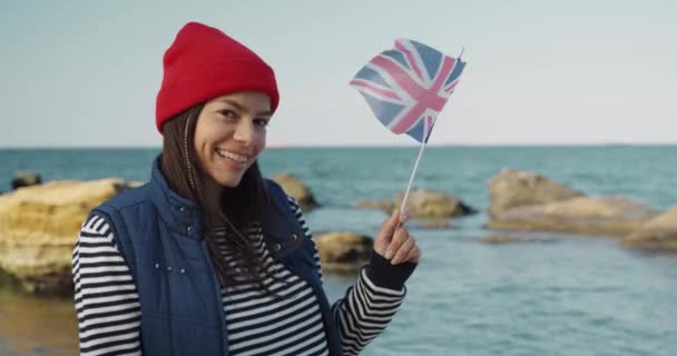 Hermosa chica turista, estudiante de intercambio sosteniendo en sus manos la bandera de Gran Bretaña y sonríe mirando a la cámara. — Vídeo de stock