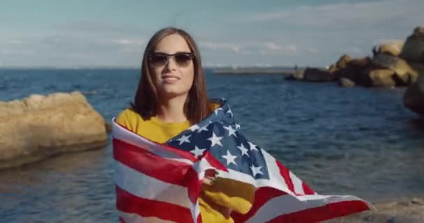 Vacker ung kvinna i gul tröja och solglasögon står på stranden insvept i flagga Amerika. — Stockvideo