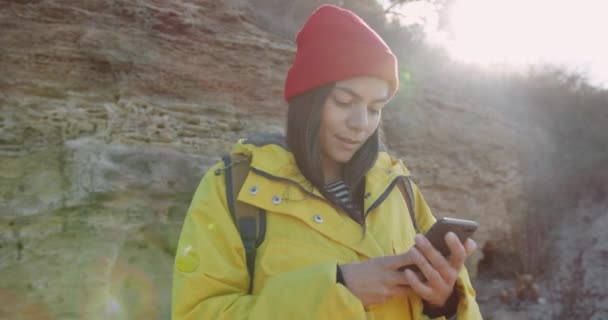 Hermosa chica caucásica turista en chaqueta amarilla de pie sobre el fondo de rocas a la luz del sol y escribe mensaje. — Vídeos de Stock