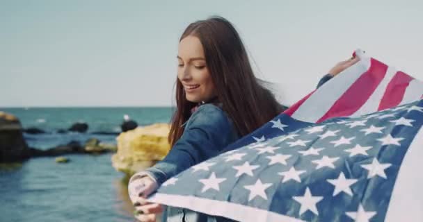 Hermosa joven de pie junto al mar desplegándose detrás de la bandera estadounidense. Estudiante de intercambio. Patriota de América. — Vídeos de Stock