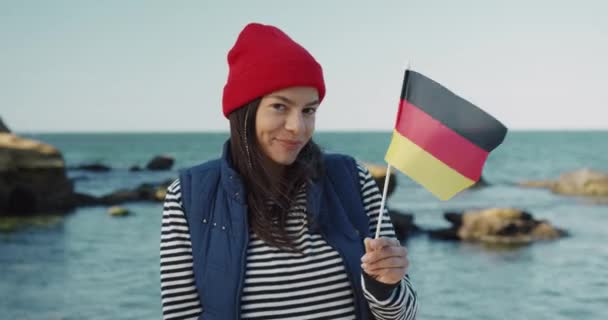 Vacker flicka turist, utbytesstudent, håller flagga i Tyskland och ler, tittar på kameran när du står på bakgrunden av havet. — Stockvideo