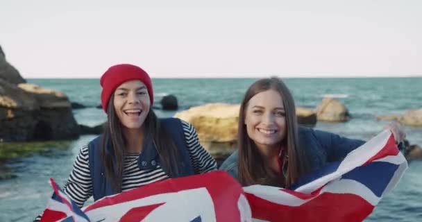 Två glada utbytesstudenter står på stranden och håller Storbritanniens flagga. Två vänner skrattar till sjöss. — Stockvideo