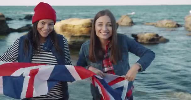 Due giovani contadine felici che camminano lungo la spiaggia con la bandiera della Gran Bretagna. Studente di scambio. Viaggiatori. — Video Stock