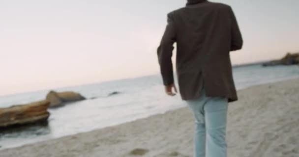 Joven hombre guapo en chaqueta de otoño runnig a lo largo de la playa y mira a su alrededor para alguien. Costa del mar. — Vídeos de Stock