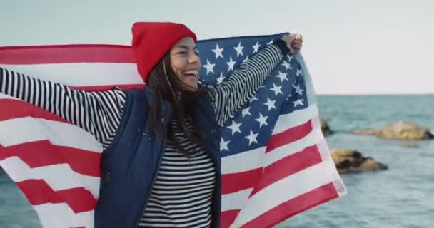 Schöne Mädchen mit rotem Hut lächelt in die Kamera und hisst die amerikanische Flagge. Glückliche Austauschstudentin mit Fahne am Meer. — Stockvideo
