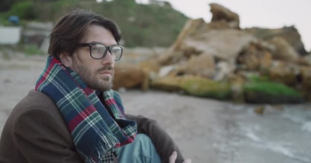 Portré jóképű fehér férfi szemüvegben és kabátban sállal ül a parton, és nézi a hullámok. — Stock videók
