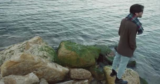 Hombre guapo y elegante en abrigo con barba caminando a lo largo del paseo marítimo y se encuentra junto al mar en piedra mirando las olas. — Vídeo de stock