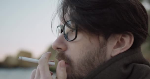 Крупный план портрета красивого кавказца с бородой и очками, сидящего на пляже и курящего сигарету. — стоковое видео