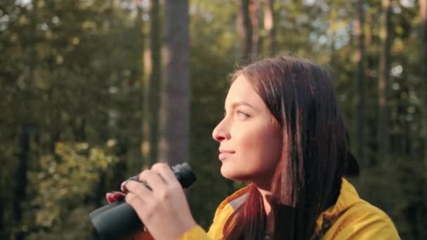 Menutup gadis cantik mengenakan jaket kuning berdiri di atas glade hutan di bawah sinar matahari dan melihat ke kejauhan menggunakan teropong. — Stok Video