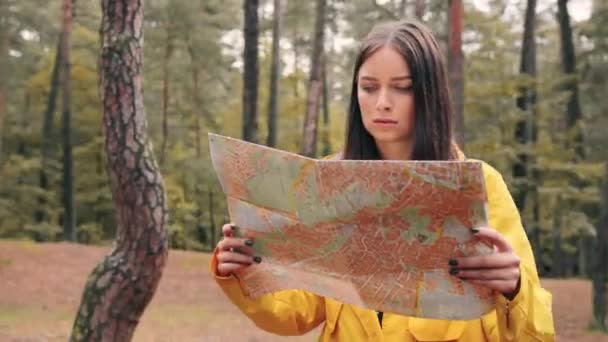 Menina branca bonita nova na jaqueta amarela que está no meio da floresta e olhando ao redor segurando confusamente mapa de papel nas mãos. — Vídeo de Stock