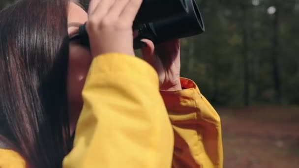 Gros plan de jeune belle femme en veste jaune debout sur la nature et regardant à travers des jumelles souriantes. Voyage à travers le pays. — Video