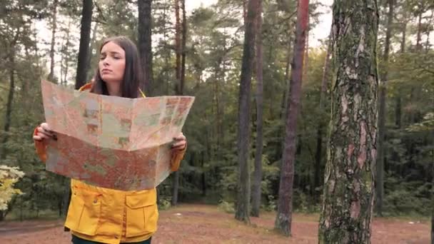 Одна дівчина в жовтій куртці стоїть серед дерев у лісі, тримаючи розгорнуту карту паперу площі, наляканий і шукає правильний шлях, щоб вийти . — стокове відео