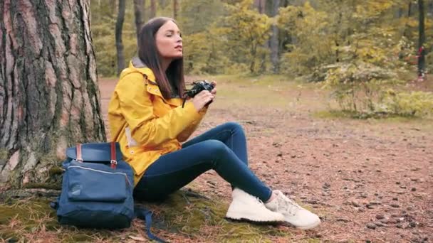 Jovem linda turista menina em casaco amarelo senta-se com mochila sozinho em madeiras sob a árvore e tira fotos na câmera. — Vídeo de Stock