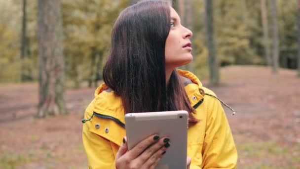 노란 재킷을 입은 소녀는 숲 속에 앉아 쉬면서 전자 타블렛을 사용하여 친구들에게 메시지를 찾아 쓰고 있다. — 비디오