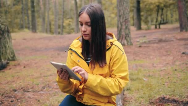 노란 자켓을 입은 소녀는 숲 속 빈터에 앉아 전자 타블렛을 사용하는 방법을 찾고 있다. — 비디오