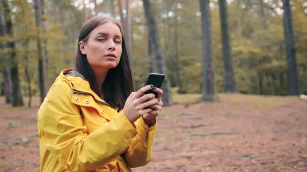Ung flicka i gul jacka sitter i skogen, koppla av, titta runt och skriva meddelanden med mobiltelefon. — Stockvideo