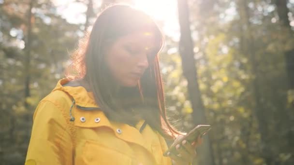 Bella ragazza solitaria in piedi nel bosco sullo sfondo della luce del sole si guarda intorno e digita il messaggio sul telefono. — Video Stock