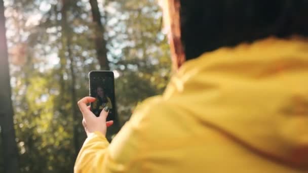 아름다운 아가씨가 숲에 서서 햇빛 아래 자연의 배경에 놓여 있는 스마트폰으로 셀카를 찍고 있습니다. 뒤에서 바라본 풍경. — 비디오