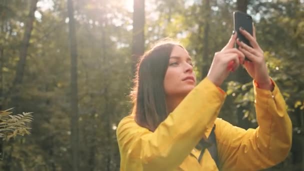 Krásná stylová dívka chodí v lese ve žlutém plášti a fotí přírodu pro sociální sítě na svém smartphonu. — Stock video