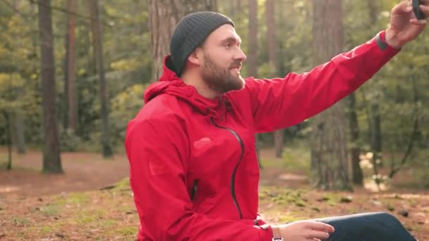 Primer plano del hombre con estilo en sombrero y chaqueta roja sentado en el bosque y hace una foto en el fondo de la naturaleza en su teléfono. — Vídeos de Stock