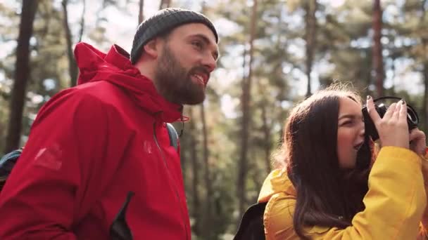 Unga glada par vandrare står i skogen på bakgrund av solljus. Vacker flicka visar foto på kameran för sin pojkvän att hon bara tog. — Stockvideo