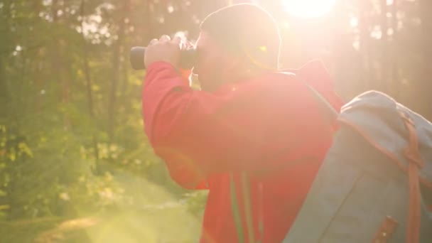 Young handsome Caucasian man in red jacket standing alone in woods in sunlight and uses binoculars to view nature. — Stock Video