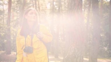 Kafkasyalı güzel fotoğrafçı kız güneş ışığında sırtında sırt çantasıyla ormanda yürür. Etrafına bakar ve kamera önünde fotoğraf çeker..