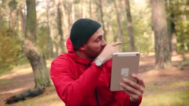 Şapkalı mutlu yürüyüşçü gündüz parkta oturuyor ve tablet kullanarak video üzerinden konuşuyor..