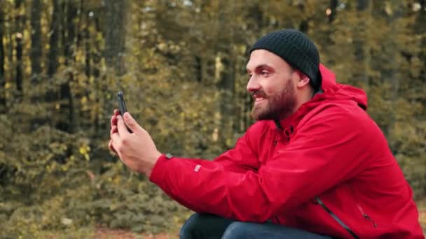 Joyeux randonneur assis dans la forêt le jour et parlant à des amis par appel vidéo à l'aide d'un smartphone. — Video