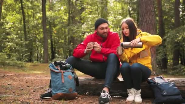 Unga par turister stannade till på stocken i skogen. Kvinna häller varmt te från termos i badtunna medan vila under resan. — Stockvideo