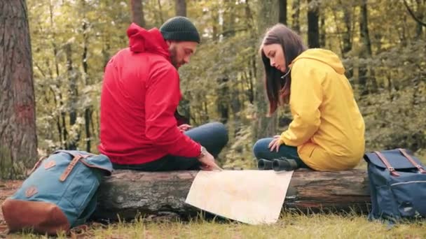 Dos turistas: hombre y mujer, sentados en el bosque en el tronco y estudiando el mapa de papel de la zona y discutiendo la siguiente ruta. — Vídeos de Stock
