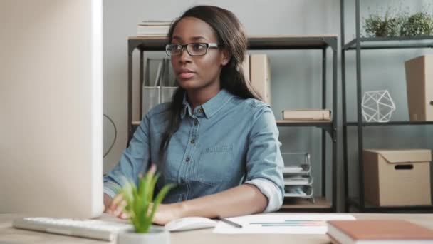 Joven chica afroamericana hermosa en gafas y camisa de mezclilla sentado en la oficina moderna trabajando en la computadora haciendo notas en documentos de papel. — Vídeo de stock