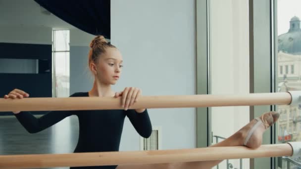 Klein meisje in zwarte sport zwembroek is oefenen terwijl leunen naar voren houden van een been op houten machine. — Stockvideo