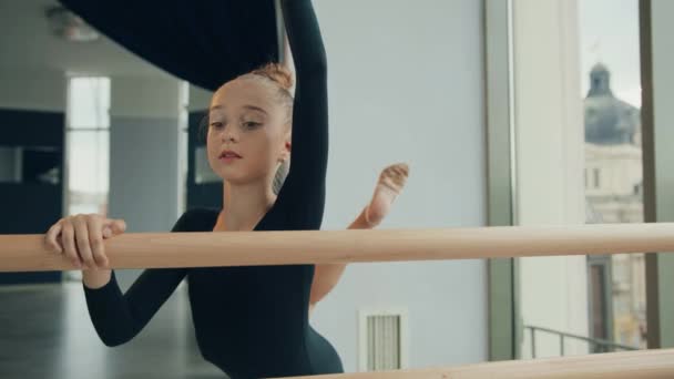 Petite fille debout dans le studio tenant par la machine à ballet et fait squats sur une jambe avec la main vers le haut. — Video