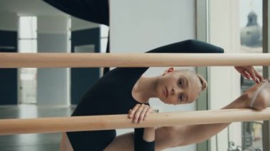 Siyah mayo giymiş şirin küçük bir kız stüdyoda duruyor. Bir bacağı bale makinesine atılmış ve kameraya bakıp gülümsüyor..