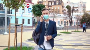 Sağlık maskeli beyaz, yakışıklı, orta yaşlı bir işadamı elinde kahve ve cep telefonuyla şehirde dolaşıyor. Yakışıklı bir adam geziniyor ve cep telefonuyla konuşuyor. Sıcak bir şeyler iç..