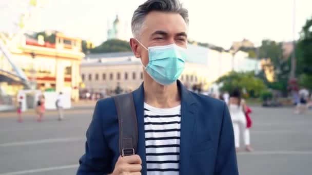 Alegre hombre guapo de mediana edad en máscara médica y bolsa caminando por la calle y mirando a la cámara. Afuera. De cerca. Un hombre de negocios guapo paseando por la ciudad en un día soleado de verano. Covid-19. — Vídeos de Stock