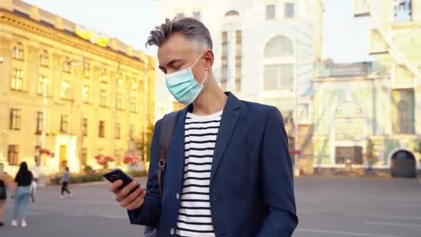 Kavkazský pohledný podnikatel v ochraně dýchacích cest na ulici a textování zpráv na mobilním telefonu. Hezký muž v masce stojící u ruského kola a poklepávající nebo posouvající se po smartphonu. — Stock video