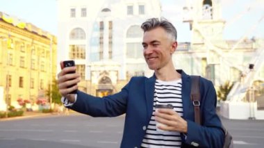 Beyaz tenli yakışıklı orta yaşlı işadamı, caddede yürüyor, kahve içiyor ve telefonda videochat ile konuşuyor. Neşeli bir adam cep telefonunda geziniyor ve görüntülü sohbet ediyor. Sıcak bir şeyler iç..
