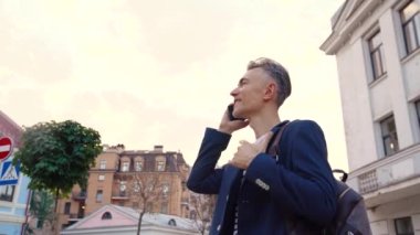 Beyaz tenli yakışıklı, neşeli orta yaşlı iş adamı caddede duruyor ve cep telefonunda gülümsüyor. Dışarıda cep telefonuyla konuşan mutlu adam. Neşeli erkek telefonda sohbet ediyor..